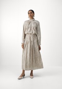 Rochie lungă, bej, cu fustă plisată și gât înalt. Prezintă un material texturat cu un model subtil. Asortată cu pantofi cu vârf ascuțit, cu spatele deschis.