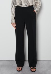 Pantalons larges noirs en tissu lisse, avec des poches latérales et une taille haute, associés à des sandales noires à lanières.