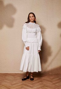 Λευκό φόρεμα με λεπτομέρειες δαντέλας και φουσκωτά μανίκια, με εφαρμοστό μπούστο και φούστα σε στρώσεις. Συνδυάζεται με καφέ παπούτσια Mary Jane.