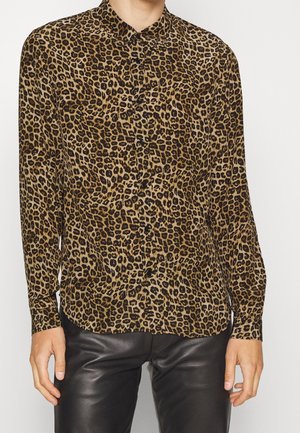 Personne portant une chemise à manches longues imprimé léopard avec un pantalon en cuir noir, debout sur un fond blanc.