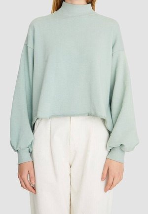 Vaalea mintunvihreä lyhyt huppari, jossa on pitkät, puffimaiset hihat ja korkea mock neck -kaulus. Pehmeä kangas ja rento leikkaus, yhdistettynä valkoisiin housuihin.