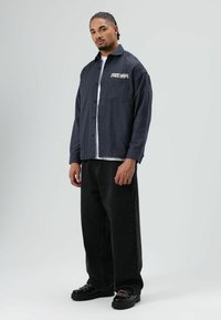 Donkerblauw geruit overhemd met knopen en witte tekst "FREE DOM", gedragen over een wit T-shirt, gecombineerd met loszittende zwarte jeans en zwarte schoenen.