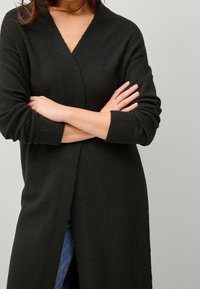 Tmavě zelený dlouhý cardigan s výstřihem do V, ribbenými rukávy a strukturovaným pletením. Přední část je otevřená, což odhaluje modré džíny pod ním.