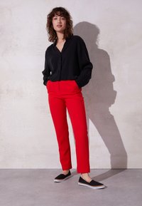 Femme debout les mains dans les poches, portant une chemise noire ample à boutons, un pantalon rouge vif et des chaussures noires sans lacets devant un mur uni.