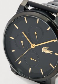 Μαύρο ρολόι χειρός με χρυσούς δείκτες και δείκτες ωρών, τρεις υποδείκτες για 24ωρη ένδειξη, μήνα και ημερομηνία, και ένα χρυσό λογότυπο κροκοδείλου στις 3 η ώρα.