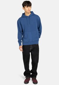 Blue cotton hoodie with a front pouch pocket and adjustable drawstring hood, paired with black loose-fit jeans and maroon footwear.