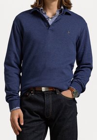 Tummansininen pitkähihaisen polo-paita, jossa on kaulus, kaksi nappia ja brodattu logo; kerrostettu valkoisen raidallisen paidan päälle ja yhdistetty tummiin farkkuihin.