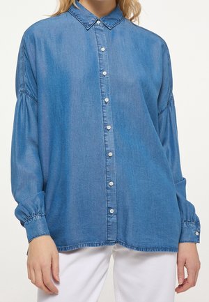 Kvinde iført en løstsiddende, langærmet blå denimskjorte med knapper og opsamlede manchetter samt hvide bukser mod en ensfarvet baggrund.