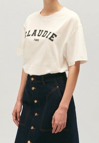 Henkilö, jolla on päällä valkoinen T-paita, jossa on teksti "CLAUDIE PARIS", sekä korkean vyötärön tummansiniset farkkuhameet, joissa on edessä kultaiset napit ja ompelureunat.