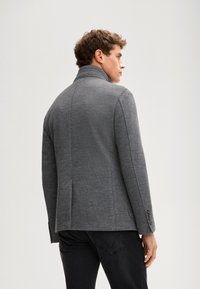 Szara kurtka z wysokim kołnierzem, dopasowanym krojem, subtelną fakturą i trzema guzikami na mankietach. Noszona na ciemnych dżinsach.