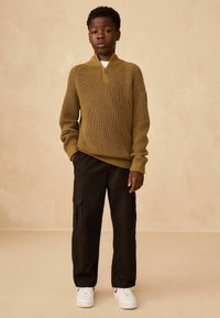 Olivgrüner, gerippter Strickpullover mit Viertelreißverschluss; kombiniert mit schwarzen Cargo-Hosen mit Seitentaschen; weiße Sneakers mit schwarzen Akzenten.