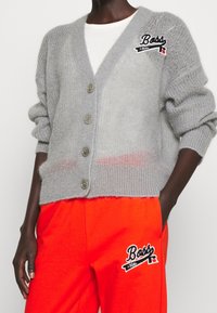 Grå, fluffig kardiagan med v-ringning, sex knappar och sidofickor. Broderad logotypdetalj på vänster bröst. Matchad med klara orange byxor.