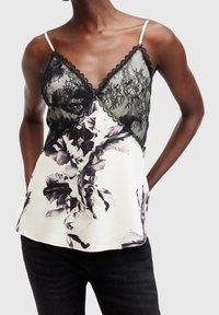 Femme portant un haut sans manches en dentelle noire et motifs floraux blancs avec des bretelles fines et un pantalon noir, debout, les mains dans le dos.