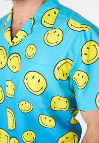 Chemise bleue à manches courtes avec un motif répétitif de visages souriants jaunes. Comprend une poche poitrine avec une étiquette cousue.