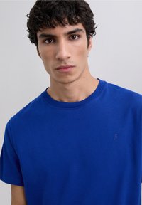 Giovane uomo con capelli ricci e scuri, che indossa una maglietta blu a girocollo semplice, che guarda direttamente nella telecamera su uno sfondo grigio chiaro.