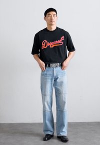 Melnā T-krekls ar sarkano "Dsquared2" logotipu, gaiši zila platai kāju džinsi ar pārlīmju detaļām un melnas kurpes. Vienkārša, monohroma estētika.