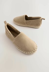 Zapatos beige de ante tipo slip-on con suela decorada con yute, con punta redondeada y un pequeño lazo trasero. Textura suave, diseño minimalista.