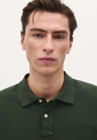Pólvora verde oscuro con cuello, que presenta una tapeta de un solo botón y un logo sutil en el pecho. Tejido texturizado.
