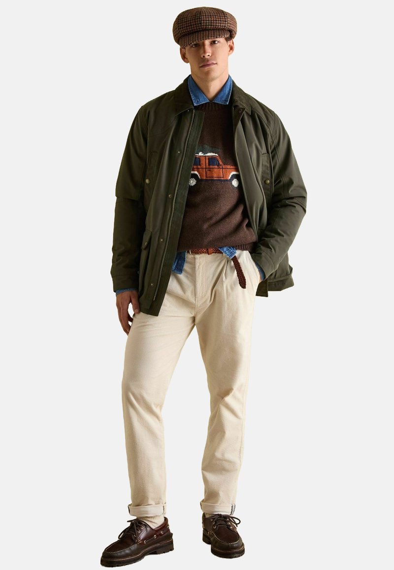 Zielona lekka kurtka, brązowy sweter z grafiką samochodu, niebieska koszula jeansowa, kremowe spodnie z kordurowej tkaniny, brązowe skórzane buty i czapka w wzory.