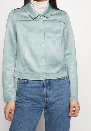 Kobieta ubrana w jasnoniebieską kurtkę na guziki na białym golfie, w połączeniu z niebieskimi dżinsami, stoi na jednolitym tle.