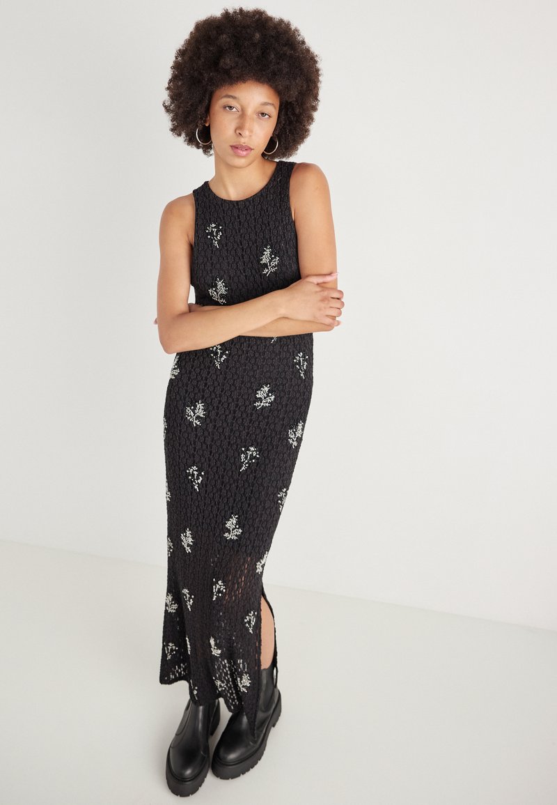 Rochie maxi neagră, fără mâneci, dintr-un material texturat, cu broderie florală albă, având o fendiră la margine și purtată cu bocanci negri masivi.