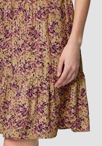 Blumenkleid mit beigem Grund, das lila und lavendelfarbene Punkte aufweist. Strukturiertes Material, geraffter Saum und geraffte Taille. Hand auf dem Stoff sichtbar.