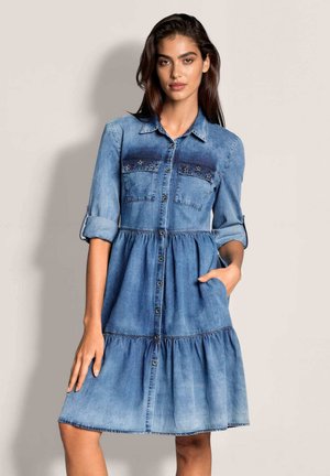 Frau mit langen dunklen Haaren, die ein blaues Denim-Kleid mit Knopfleiste, hochgekrempelten Ärmeln und Taschen trägt, steht vor einem einfarbigen Hintergrund.