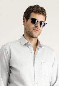 Camicia button-up grigia chiara con colletto, dotata di taschino sul petto e dettagli di cucitura sottili, indossata con occhiali da sole neri.