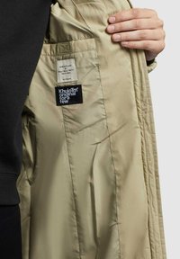 Bežs mēteles iekšpuse ar gludu tekstūru, kreisajā pusē ir divas etiķetes - viena ar logo, otra ar rūpības norādi.