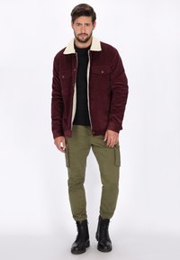 Bordeauxfarvet fløjlsjakke med en cremefarvet shearlingkrave, to brystlommer, parret med olivengrønne cargo-bukser og sorte snørebåndsstøvler.