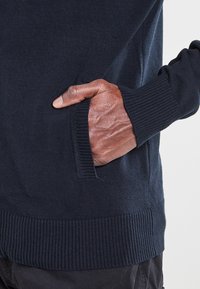 Pull bleu marine avec poignets et ourlet côtelés, doté d'une poche latérale. Tissu doux et texturé visible le long du bord de la poche.