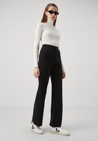 Svarte høytlivbukser med glidelås, parret med en hvit ribbestrikket turtleneck og hvite sneakers med svarte detaljer.