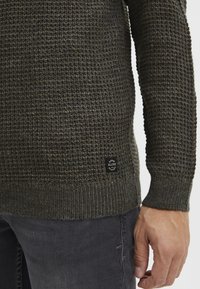 Dunkelgrüner Strickpullover mit strukturiertem Muster, gerippten Bündchen und Saum. Verfügt über ein rundes Logopatch auf der unteren linken Seite.