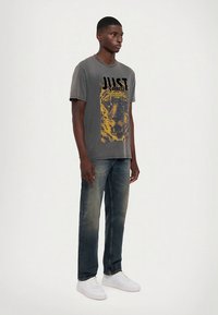 Grå bomulls t-skjorte med en grafikk av en tiger og svart tekst på forsiden, kombinert med falmede blå jeans og hvite joggesko.