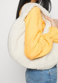Bolso de crema redondo y esponjoso hecho de material texturizado, con bordes suaves. El bolso lo sostiene una persona que lleva una blusa amarilla arrugada.