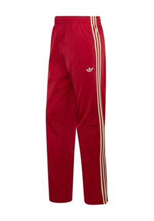 Czerwone dresowe spodnie Adidas z beżowymi potrójnymi paskami po bokach oraz kieszenią na zamek po prawej stronie biodra.