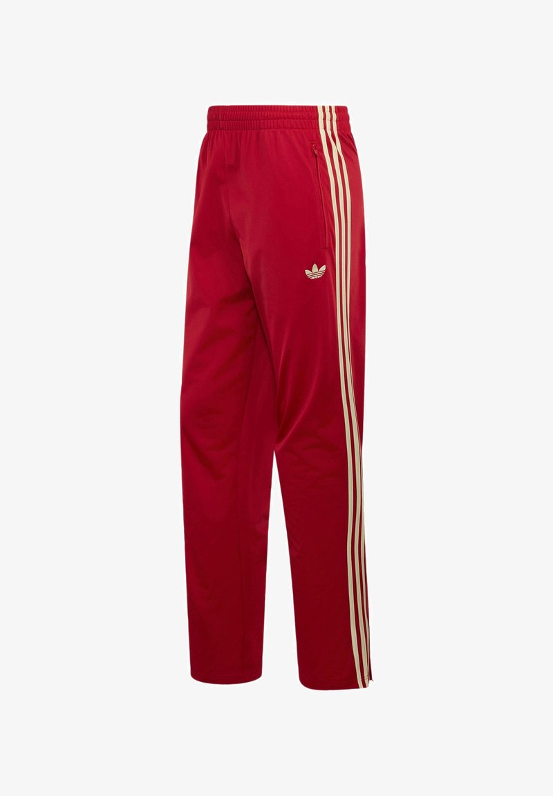 Røde Adidas træningsbukser med beige tredobbelte striber ned langs siderne og en lynlåslomme på højre hofte.