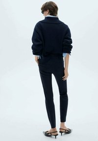 Námořnický oversized pletený svetr s vysokým límcem, v kombinaci s slim-fit tmavými kalhotami a černými sandály na podpatku. Tkanina vypadá měkce a texturovaně.