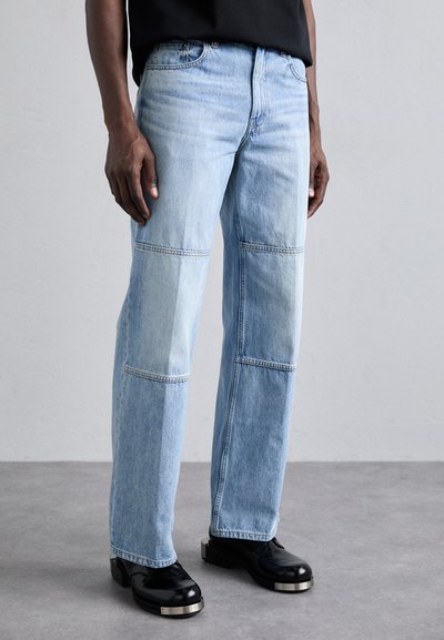 Light blue denim jeans with a patchwork design, featuring two-tone panels and contrasting stitching. Paired with black shoes with metallic accents.