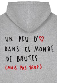 Sivá mikina s kapucňou na šnúrku. Na prednej strane je vyšívaný text čiernou a červenou farbou: "UN PEU D'♥ DANS CE MONDE DE BRUTES (MAIS PAS TROP)".