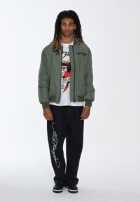 Giacca bomber verde oliva, T-shirt bianca con design di tatuaggio, pantaloni di denim neri con scritta bianca e scarpe nere con suola chiara.