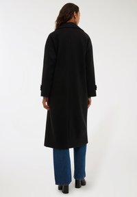 Czarny wełniany płaszcz o długiej sylwetce, szerokich klapach i guzikach na rękawach, zestawiony z niebieskimi dżinsami i czarnymi botkami na obcasie.