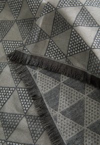 Tkanina z geometrycznym wzorem przeplatających się czarnych i szarych trójkątów. Krawędzie są ozdobione frędzlami dla tekstury, z różnorodnymi splotami tkaniny.