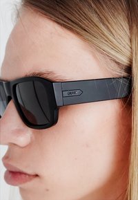Gafas de sol negras con montura rectangular, lentes oscuros y un acabado mate. Las varillas presentan un sutil detalle del logo en el costado.