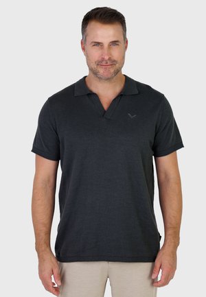 Mężczyzna o krótkich brązowych włosach i jasnej brodzie, ubrany w czarną koszulkę polo z krótkim rękawem i beżowe spodnie, stojący na jednolitym tle.