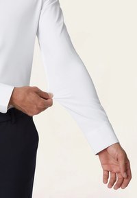 Witte lange mouw overhemd met een fitted ontwerp; gladde textuur; manchetten met één knop; hand die de mouw aanpast. Achtergrond is een lichte neutrale kleur.