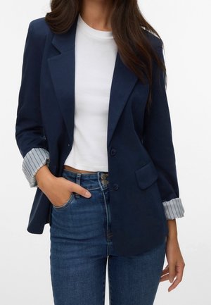 Kvinde iført en navy blazer med stribede manchetter, en hvid cropped t-shirt og højttaljede blå jeans med to synlige knapper.