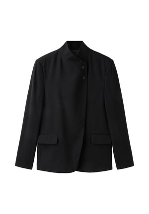 Femme portant une veste noire boutonnée, un jean large déchiré et des chaussures noires, tenant un petit sac à main noir sur un fond gris.