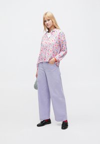 Blusa rosa con estampado floral colorido y cierre de botones al frente, combinada con pantalones de pana de pierna ancha en tono lila claro. Zapatos negros y calcetines rojos.