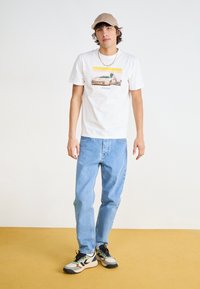 White cotton t-shirt with graphic illustration, blue denim jeans, and multicolored sneakers. Beige cap and chain necklace. Yellow background.